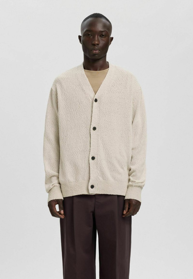 Homme à la peau foncée portant un cardigan boutonné beige texturé par-dessus une chemise beige et un pantalon marron foncé, debout devant un fond clair uni.