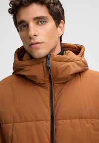 Giacca imbottita in un caldo marrone, con colletto alto, cappuccio e chiusura con zip. Tessuto liscio con dettagli imbottiti cuciti.