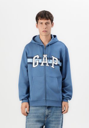 Jeune homme portant un sweat à capuche zippé bleu avec le logo "GAP" et un jean bleu, se tenant devant un fond blanc uni.