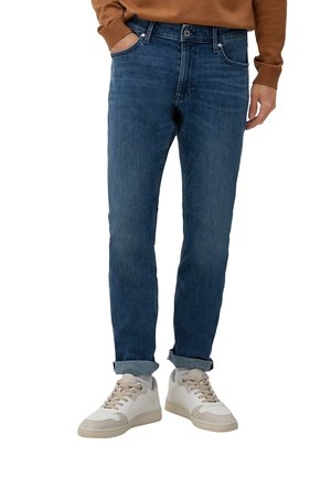Homme portant un jean bleu retroussé, des baskets blanches et une chemise marron à manches longues, debout avec une main dans la poche.