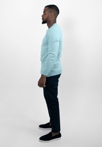 Lichtblauwe trui, slim-fit donkere broek en zwarte instapschoenen. Model posed in profiel tegen een effen achtergrond.