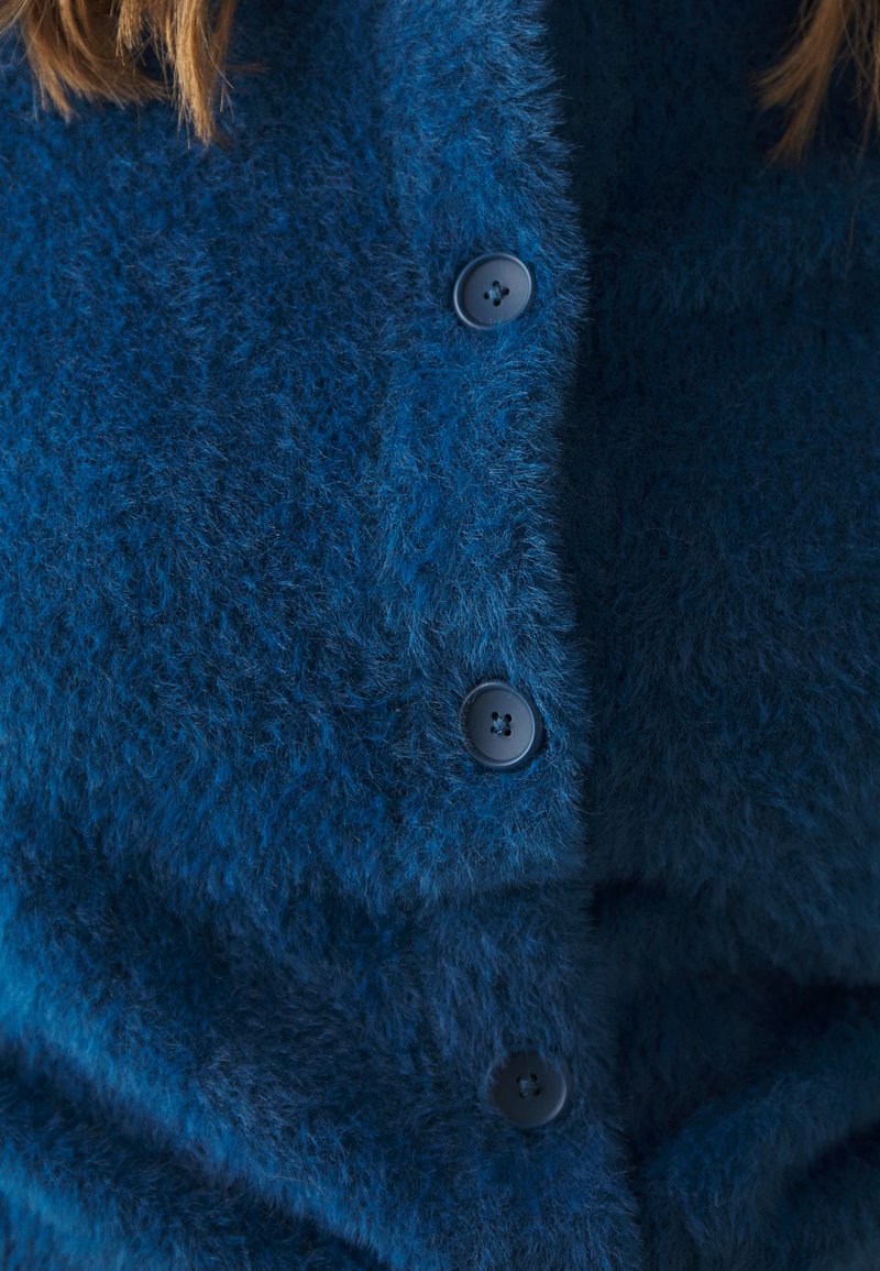 Cardigan bluiccio e morbido con una texture soffice, bottoni neri arrotondati e un design con bottoni. Ingrandimento del dettaglio frontale e del tessuto.