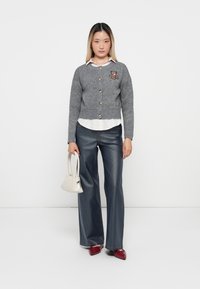 Cardigan gris avec des boutons et une broderie d'ours en peluche, superposé sur une chemise blanche, associé à un jean large et des chaussures rouges.