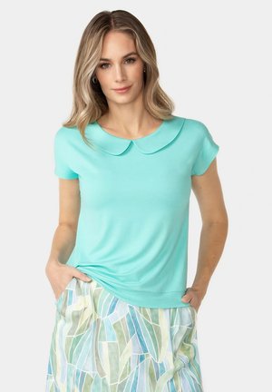 Frau mit langen blonden Haaren, die ein türkisfarbenes Kurzarmoberteil mit Peter-Pan-Kragen und einen gemusterten Pastellrock trägt, die Hände in den Taschen.