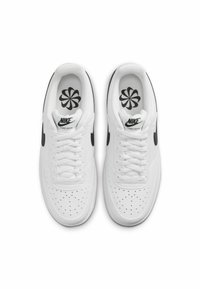 Baskets Nike blanches avec une boîte à orteils perforée, un logo Swoosh noir et des semelles intérieures à motifs, présentant une tige en cuir lisse et des lacets plats.