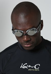 Zwarte zwembril met zilveren gespiegeld lenzen en verstelbare banden, gedragen door een persoon in een aansluitend zwart zwemtop met logo details.