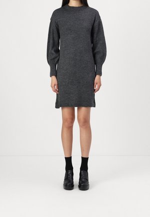 Femme portant une robe en maille gris foncé à manches bouffantes longues, des chaussettes noires et des chaussures à plateforme noires, debout devant un fond uni.