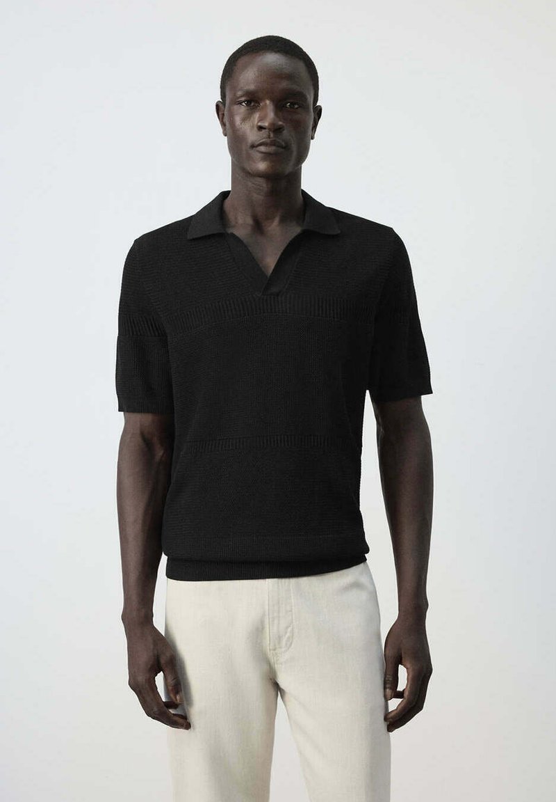 Homme debout face à l'avant, portant un polo en maille noir à manches courtes et un pantalon beige clair sur un fond uni.