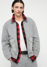 Jachetă din denim gri cu două buzunare frontale, purtată peste o cămașă din flanel cu carouri roșii și negre. Tricou alb dedesubt, curea neagră vizibilă.