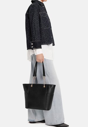 Osoba drží černou koženou tašku na rameno s řetízkovými a koženými úchyty, oblečená v černé bundě s vzorem, bílém tričku a světle modrých džínech s širokými nohavicemi.