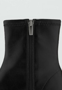 Botín de cuero negro hasta el tobillo con una cremallera metálica prominente en la parte delantera, que cuenta con detalles de costura a lo largo del borde superior y una textura suave.