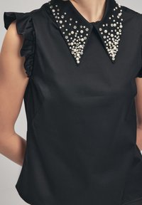 Top nero con maniche a volant e colletto a punta, decorato con perle bianche sparse e strass trasparenti, con una texture di tessuto liscia.