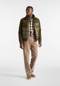 Giacca verde oliva con tasche con zip, indossata sopra una camicia a quadri gialla, blu navy e arancione, abbinata a pantaloni in corduroy beige e scarpe marroni.