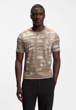 Ung mand iført en beige kortærmet mønstret skjorte og sorte bukser, stående mod en ensfarvet hvid baggrund.