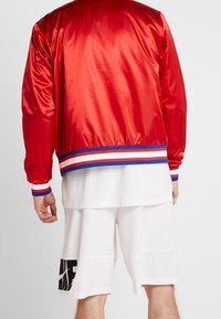 Rote Satin-Bomberjacke mit gestreiften blauen und weißen Akzenten, getragen über einem weißen Netzshirt und weißen Shorts mit einem schwarzen Grafikdruck.