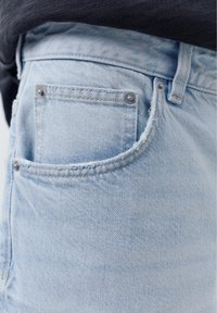 Lys blå denim jeans baglomme med metalnitter og synlige sømme, båret med en mørk skjorte stukket ind.