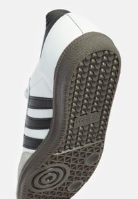 Zapatilla blanca con rayas negras; suela de goma texturizada con agarre en patrón; parte superior de cuero sintético con acabado suave y acentos de ante.
