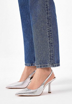 Ezüst színű fémes slingback magassarkú cipők, hegyes orral, texturált felülettel és vékony sarkú, kék egyenes szárú farmernadrággal párosítva.