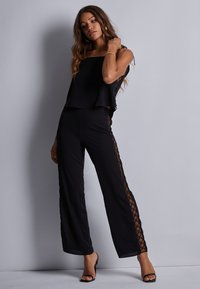 Jumpsuit noir avec un haut superposé, une coupe à jambes larges et des panneaux latéraux transparents avec un motif en diamant. Des bretelles ajustables complètent le look.