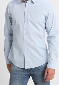 Hellblaues, langärmliges Hemd aus glattem Baumwollstoff mit spitzem Kragen, geradem Saum und weißen Knöpfen. Kombiniert mit Jeans.