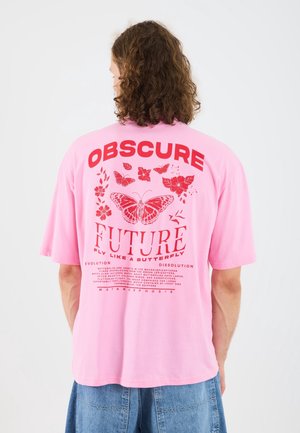 Personne aux cheveux bouclés porte un t-shirt rose avec des graphismes rouges de papillons et de fleurs, et un texte indiquant "OBSCURE FUTURE Fly Like a Butterfly."