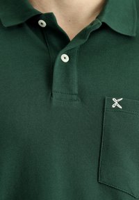 Polo verde scuro con motivo testurizzato; presenta un colletto, chiusura con bottoni e un piccolo ricamo bianco sulla tasca sul petto.