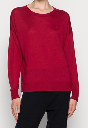 Rode trui met lange mouwen en een ronde hals, met kleine geborduurde initialen op de borst, gedragen met een zwarte broek.