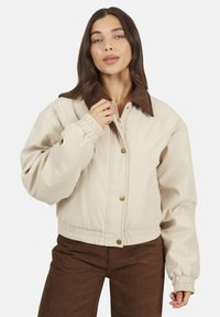 Veste cropped beige avec col en velours côtelé marron, dotée de manches longues, de poignets élastiques et de boutons-pression dorés sur le devant.