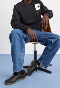Sudadera negra con logo en el lado izquierdo del pecho, jeans de mezclilla azules de corte holgado y zapatillas negras con suela de goma, sentado en una silla de madera.