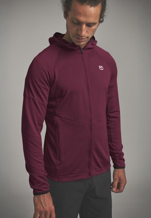 Mann trägt eine bordeauxrote Kapuzenjacke mit Reißverschluss, langen Ärmeln und Seitentaschen, schaut nach unten vor einem einfarbigen Hintergrund.