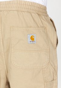 Beige cargo pants with an elastic waistband, side pockets, and a Carhartt logo patch in yellow on a square grey background.