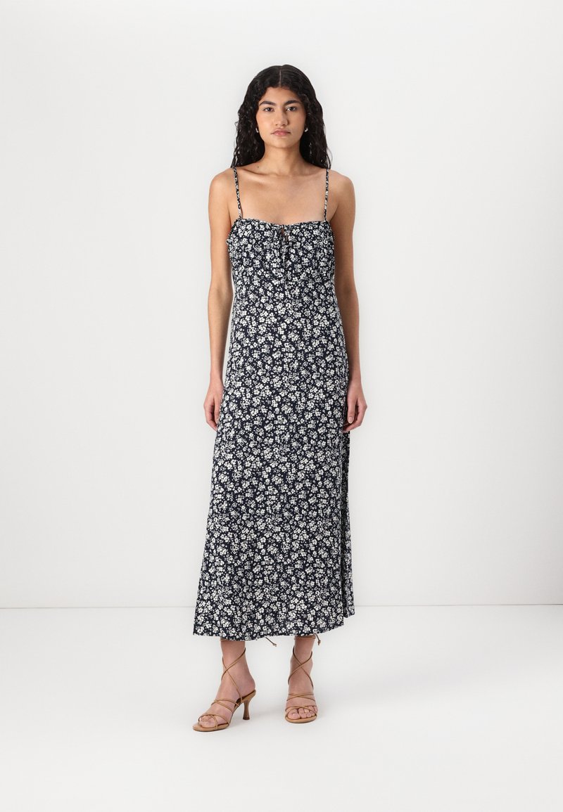 Rochie maxi floral de culoare albastru marin cu bretele subțiri, cu un corset plisat, material fluid și fante pe laterale. Poartă cu sandale cu toc de culoare bej.
