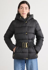 Zwarte gewatteerde jas met capuchon, geribbelde tailleband, gouden hardware en horizontale bies. Voorzien van zijzakken en een glanzende afwerking.