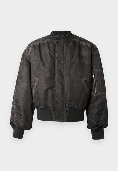Črna bomber jakna z gladko, teksturirano površino, rebrastimi manšetami in obrobom, z zadrgo žepom na rokavu in minimalističnim dizajnom.