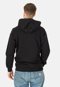 Schwarzer Kapuzenpullover mit langen Ärmeln, lockerer Passform und geripptem Saum. Tragen mit hellblauen Jeans.