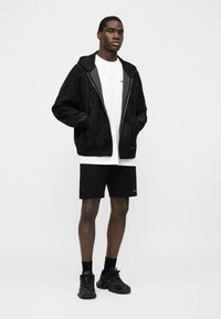 Tânăr purtând o hanorac negru cu fermoar și textură, tricou alb, pantaloni scurți negri, șosete negre și adidași negri, stând cu mâinile în buzunare.