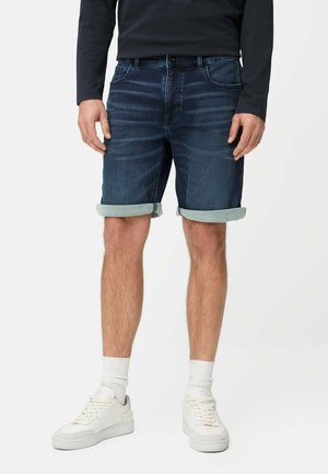 Man draagt donkere spijkershorts met opgerolde pijpen, witte sneakers, witte sokken en een zwarte longsleeve, staand tegen een effen achtergrond.