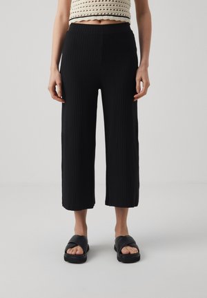 Femme portant un pantalon court côtelé noir, un haut court en crochet crème et des sandales plates noires devant un fond gris uni.