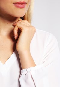 Donna con pelle chiara e labbra rosa appoggia il mento sulla mano, indossando una camicia bianca a maniche lunghe con polsini abbottonati, davanti a uno sfondo neutro.