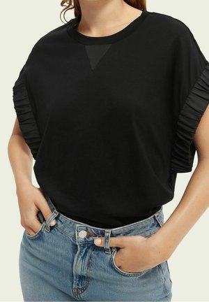 Femme portant un haut noir à manches courtes avec des détails froncés sur les manches et un jean bleu, les mains dans les poches avant.