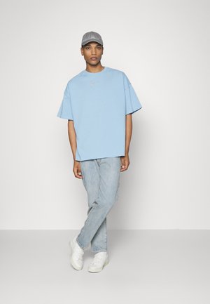 Tricou oversized de culoare albastru deschis, cu guler rotund, asociat cu blugi decolorați și adidași albi. Include o șapcă gri. Design simplu.