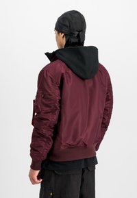 Mies, joka käyttää mustaa duragia ja kerroksellista vaatetusta mustan hupparin alla maroon-värisen bomber-takin kanssa, seisoo valkoista taustaa vasten.