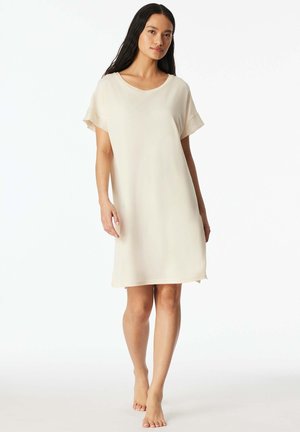 Woman with long dark hair wearing a loose, short-sleeve, knee-length cream dress, standing barefoot on a plain white background.