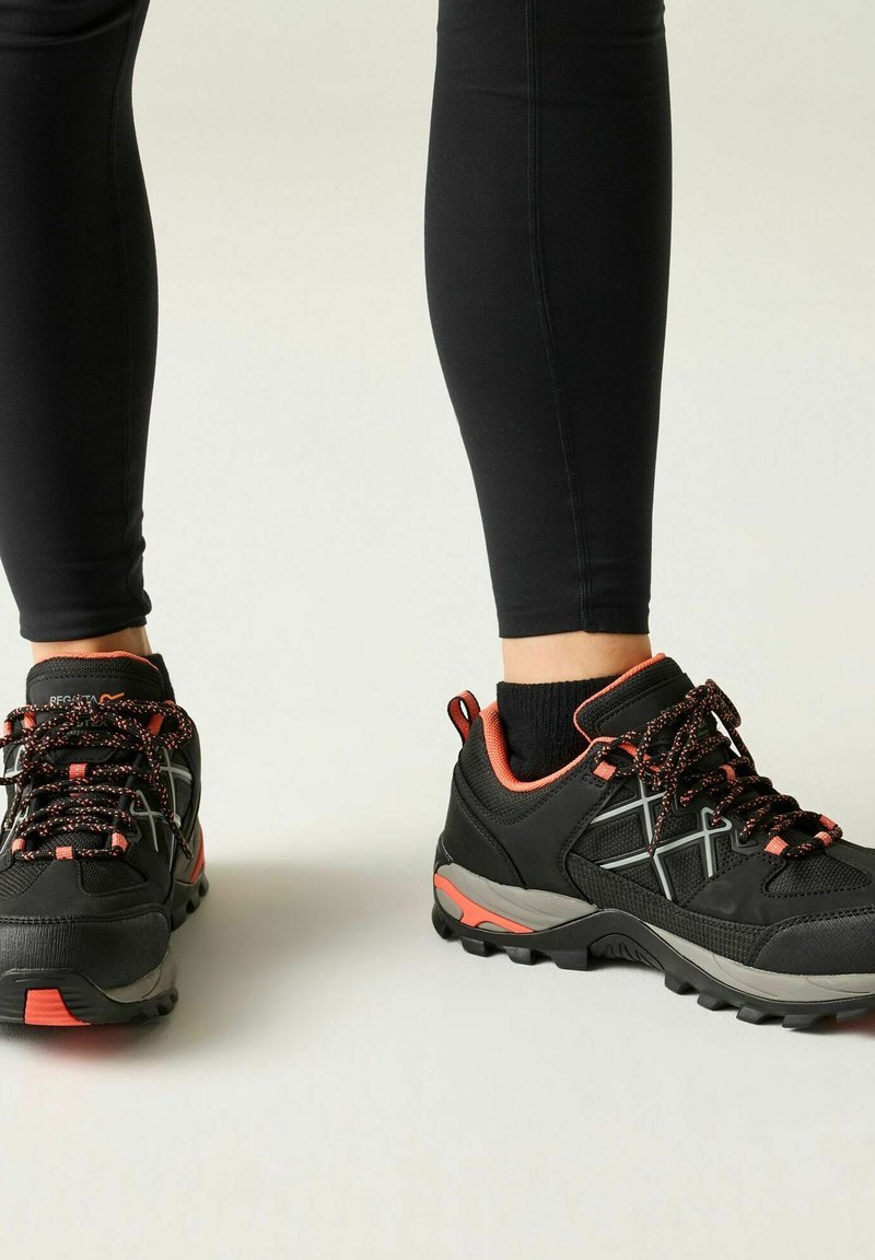 Osoba nosząca czarne i pomarańczowe buty trekkingowe oraz czarne legginsy, stojąca na jasnej powierzchni.