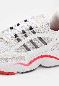 Bílá sportovní bota s horní částí z meshového materiálu, šedými detaily, třemi černými pruhy a texturovanou podrážkou s červeným pruhem na patě.