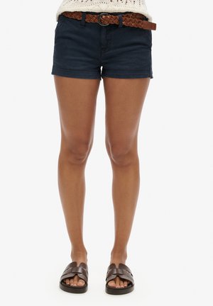 Shorts bleu marine foncé en coton avec une ceinture tissée marron. Les shorts ont une coupe décontractée et sont associés à des sandales noires plates.