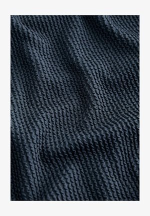 Struktūruotas, tamsiai mėlynas audinys su bangų raštu, kuris turi pakeltas briaunas, demonstruojantis švelnią, megztą išvaizdą.