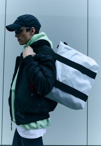 Hvit totebag med svarte stropper, blankt materiale, rektangulær form, båret på skulderen. Modell i svart bomberjakke og grønn hettegenser.