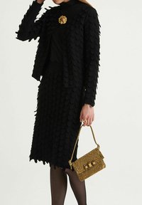 Veste noire et jupe avec une texture superposée rappelant des feuilles, associées à un sac à main doré tissé avec des accents circulaires.