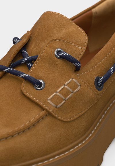 Chaussure bateau en daim beige avec lacets bleu marine et blancs, détails cousus et semelle épaisse en caoutchouc.